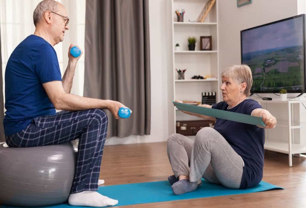 Aged people doing fitness together at home using dumbbells, swiss ball and elastic band. Old person healthy lifestyle exercise at home, workout and training, sport activity at home on yoga mat.