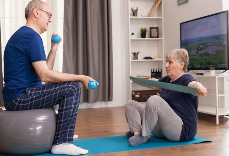 Aged people doing fitness together at home using dumbbells, swiss ball and elastic band. Old person healthy lifestyle exercise at home, workout and training, sport activity at home on yoga mat.