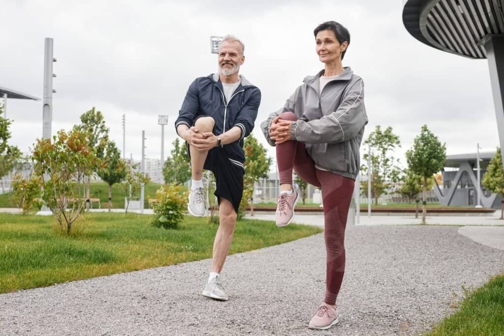 Full shot of smiling senior man and woman doing standing knee hugs, warming up and getting ready to run jogging at morning city park with gray sky