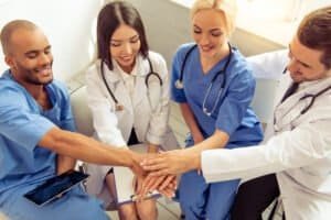 Medical doctors of different nationalities and genders are holding hands together and smiling, sitting in office