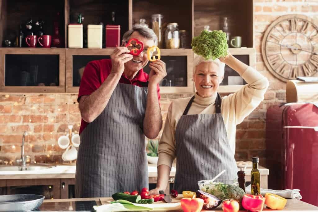 A happy senior couple, wearing aprons, are cooking together in their kitchen. The woman is holding a wooden spoon with a taste of food on it, while the man is smiling as she tastes it.