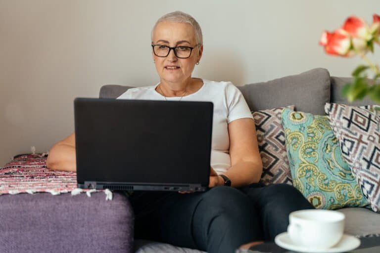 Cheerful older woman in white shirt smiles while using laptop on sofa. Relaxed mood, working or browsing online. Modern home vibe, light interior, positive senior lifestyle.